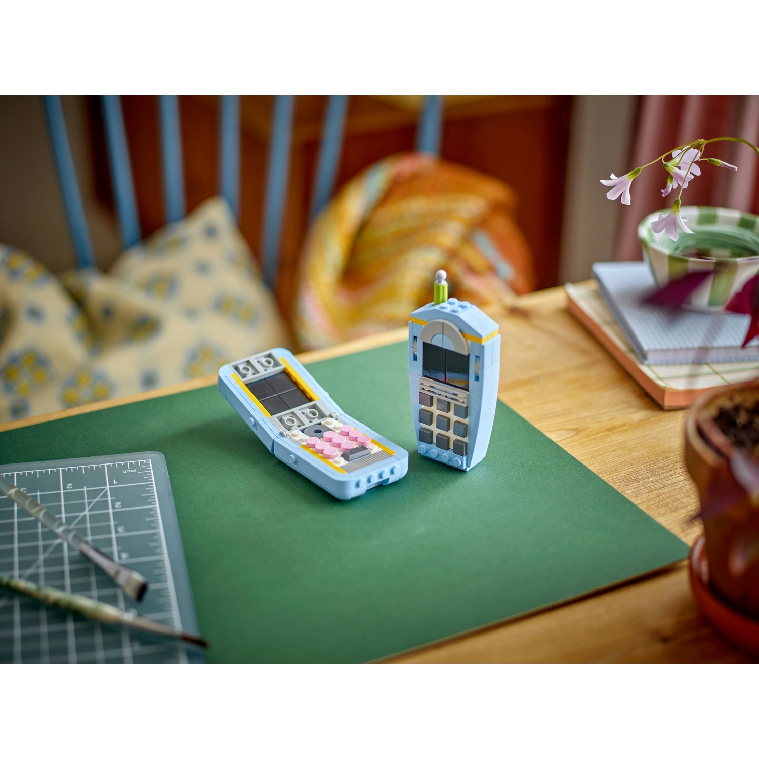 Due telefoni Lego su una superficie verde con pennelli e un tappetino da taglio. Fiori, una ciotola e una sedia sullo sfondo.