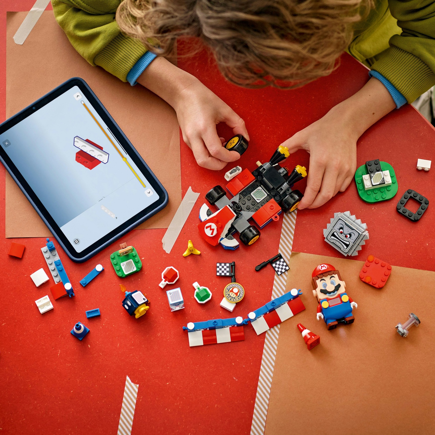 Un bambino costruisce un kart Lego su un tavolo rosso. Un tablet mostra le istruzioni. Pezzi Lego, la figura di Mario e Thwomp sono sparsi.