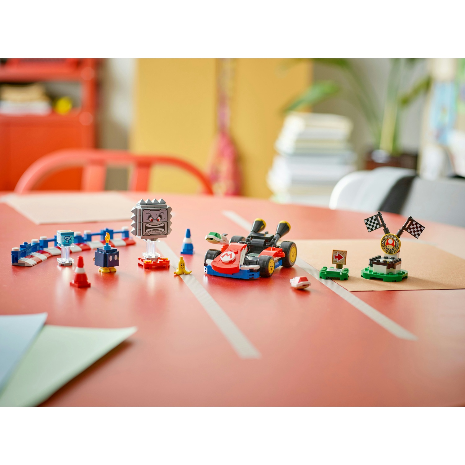 Set Lego Mario kart su un tavolo rosso. Include kart, ostacoli e traguardo. Sfondo con stanza sfocata.