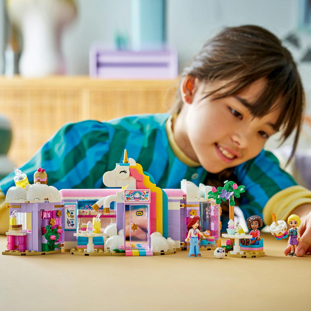 Ein Mädchen lächelt über einem LEGO-Set mit Einhorn-Motiv. Das Set enthält Gebäude, Minifiguren und Tische. Helle Farben werden verwendet.