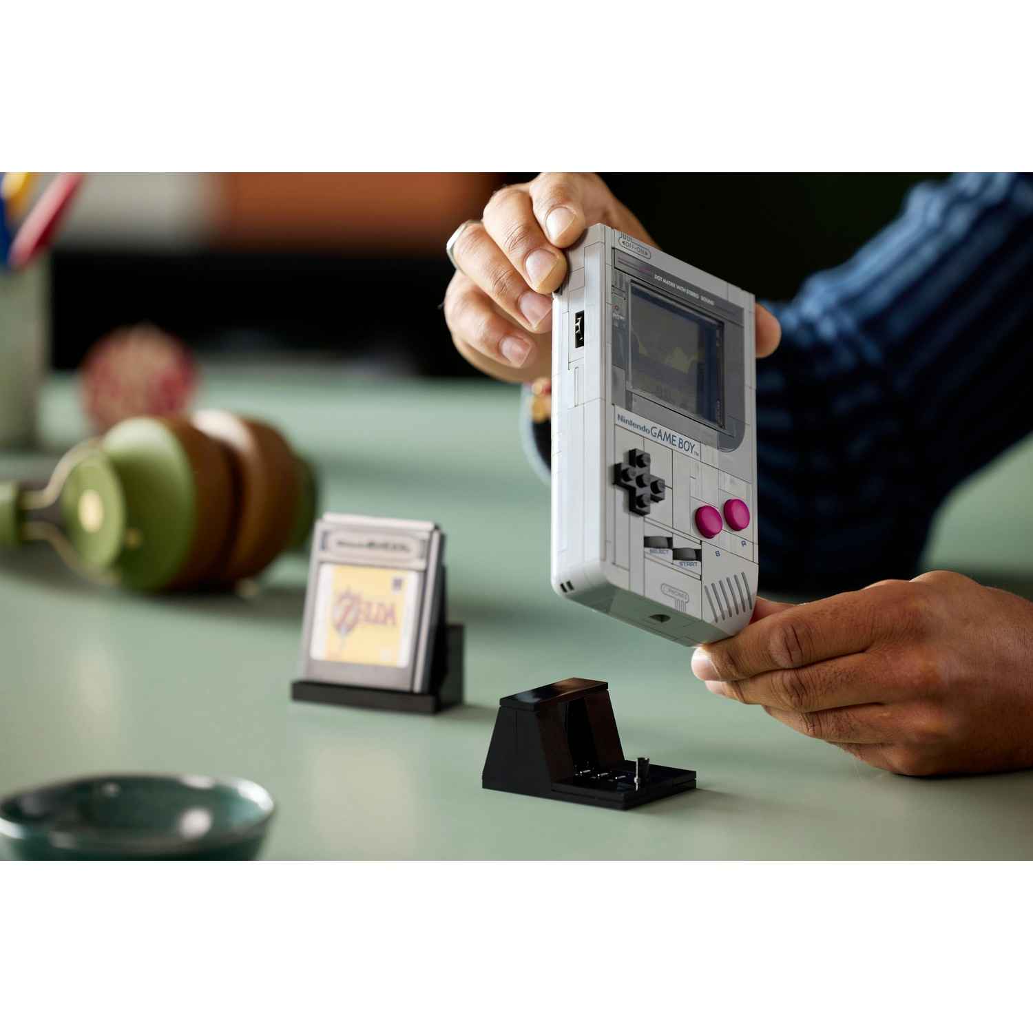 Una persona tiene in mano un modello bianco di Game Boy. Accanto, una cartuccia di gioco, una base di ricarica e cuffie su una superficie verde.