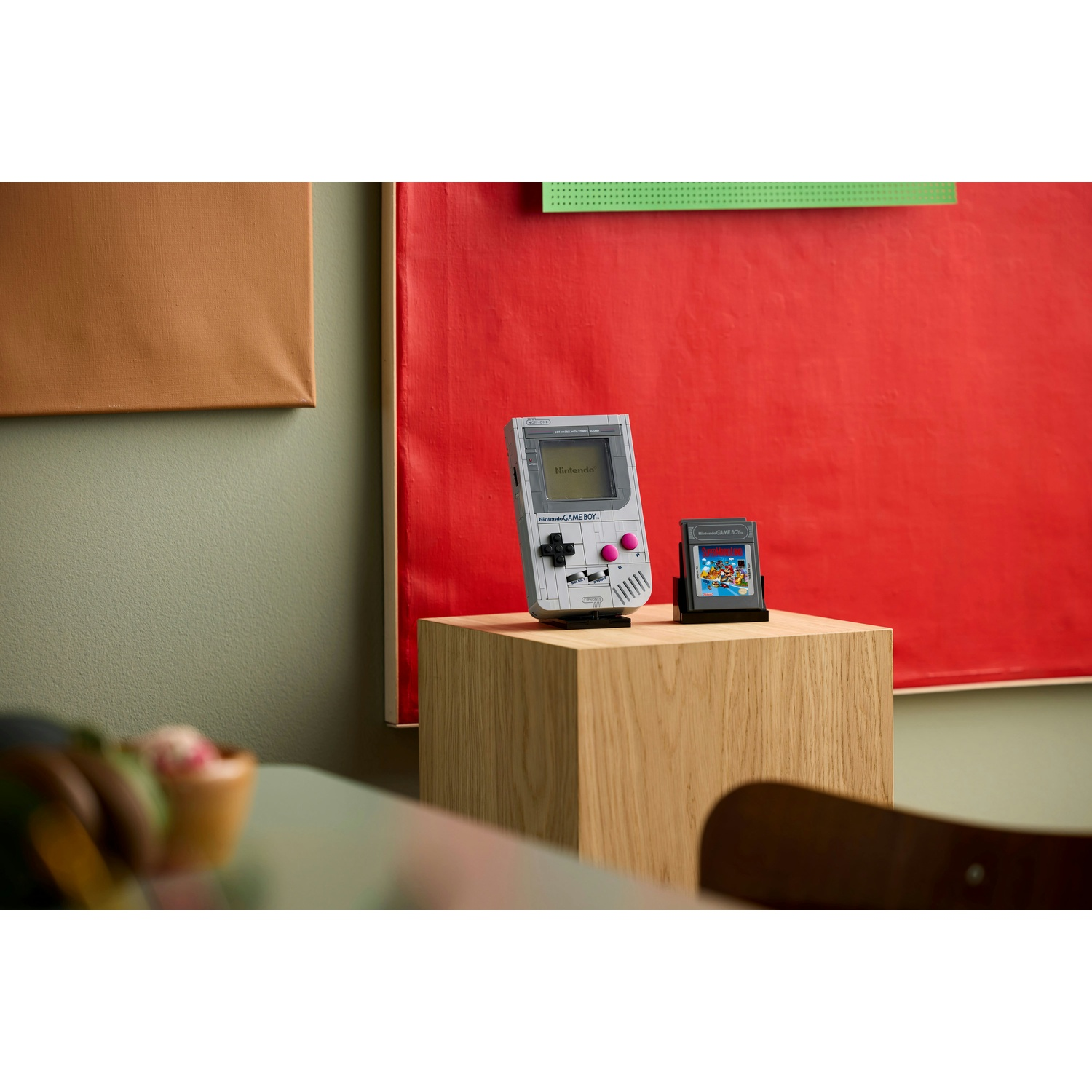 Un Game Boy bianco e grigio e una cartuccia sono su un cubo di legno, contro un muro rosso. A sinistra c'è una tela marrone.