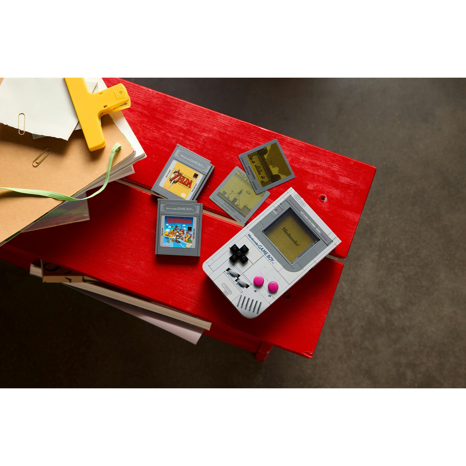 Una Nintendo Game Boy bianca, cartucce di gioco e articoli di cancelleria su un tavolo rosso. Lo sfondo è un pavimento grigio.