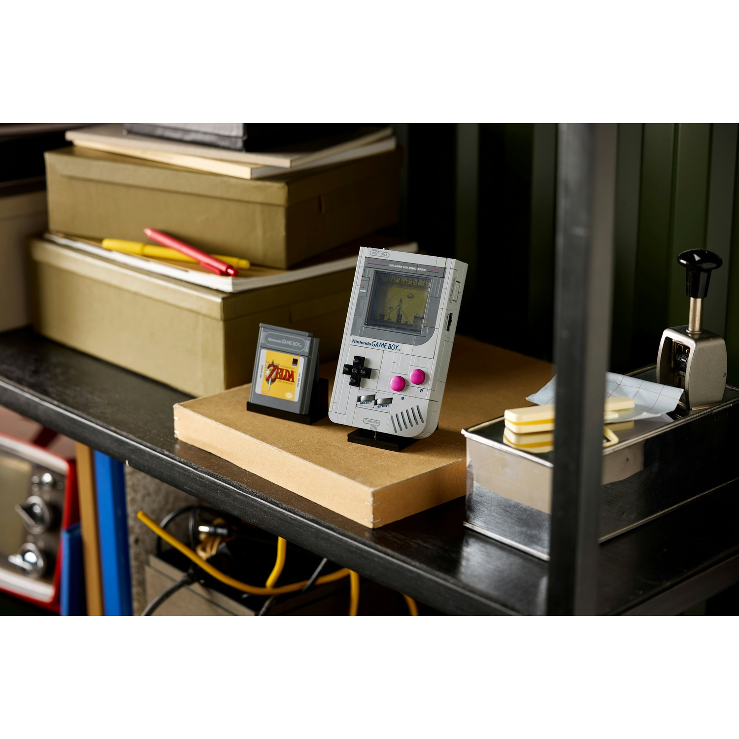 Un Game Boy Lego e una cartuccia di Zelda sono su uno scaffale. Una cucitrice e delle scatole sono sullo sfondo. Le penne poggiano su una scatola.