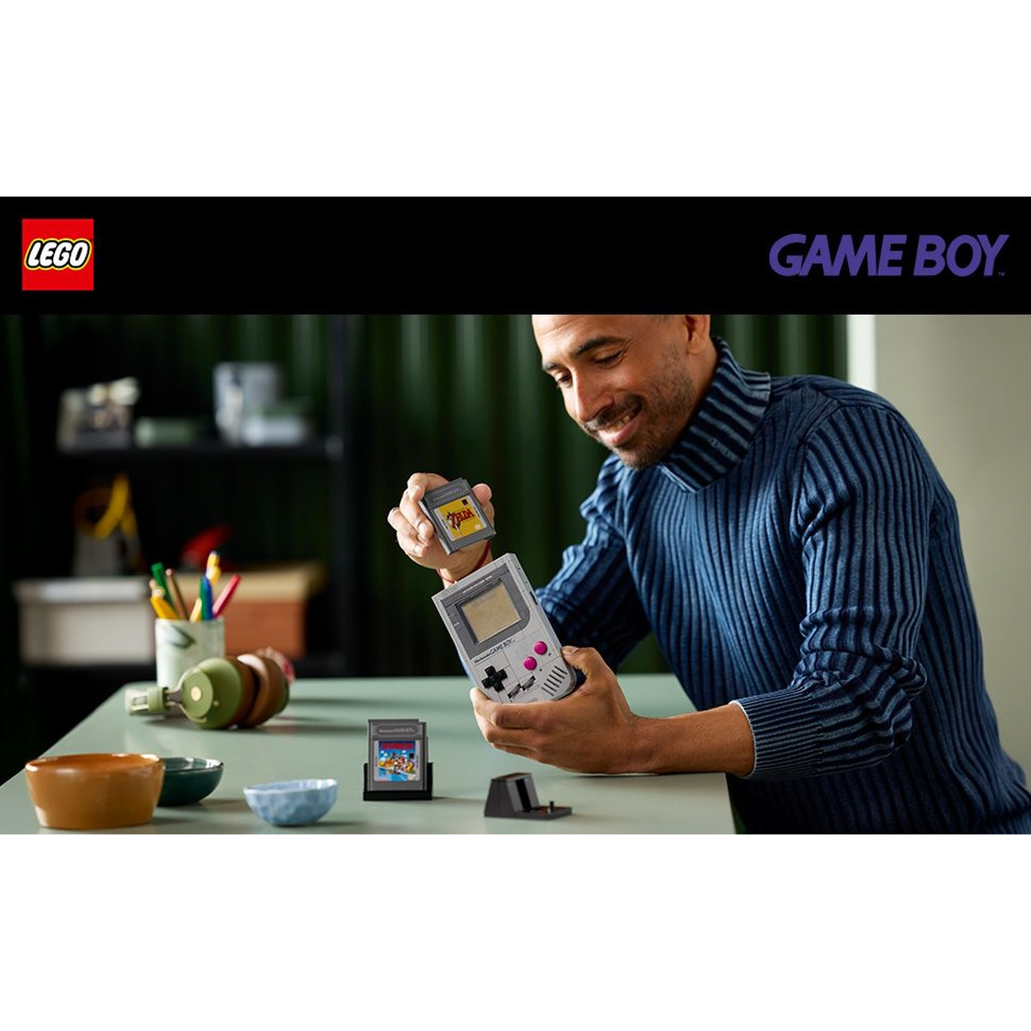 Uomo che tiene in mano un Game Boy e una cartuccia Lego. Il tavolo ha ciotole, cuffie e penne. I loghi Lego e Game Boy sono visibili.