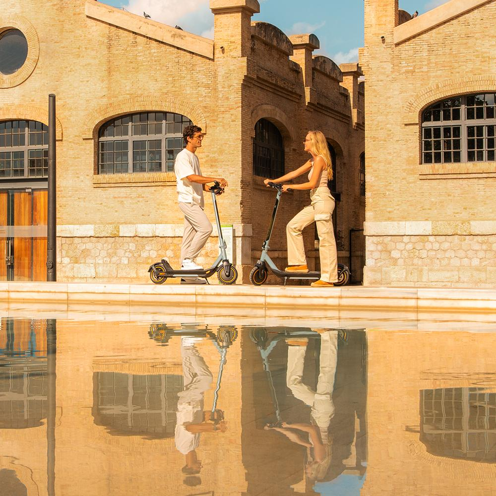 Un hombre y una mujer en patinetes frente a un edificio de ladrillo, reflejados en el agua. Cielo azul.
