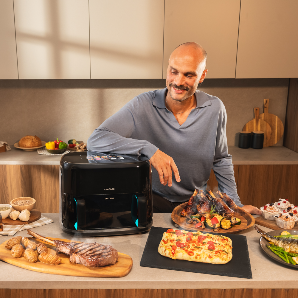 Un hombre sonríe cerca de una freidora de aire negra con alimentos como pizza, bistec y verduras en el mostrador. Fondo de cocina.