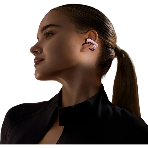 Una mujer usa auriculares rosas. Tiene cabello rubio en una cola de caballo y viste una blusa negra. Toma de perfil sobre blanco.