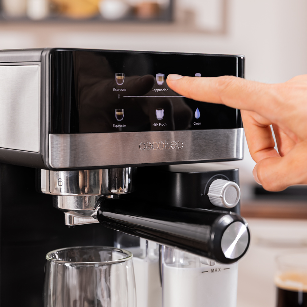 Una mano presiona el botón 'Capuchino' en una cafetera negra y plateada. Hay un vaso y recipientes de leche al frente.