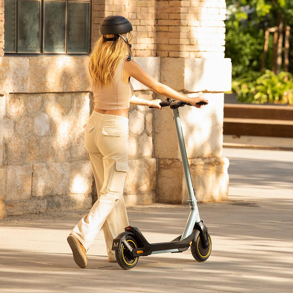 Una mujer con ropa beige y casco camina con un patinete eléctrico. El fondo muestra una pared de ladrillo y vegetación.