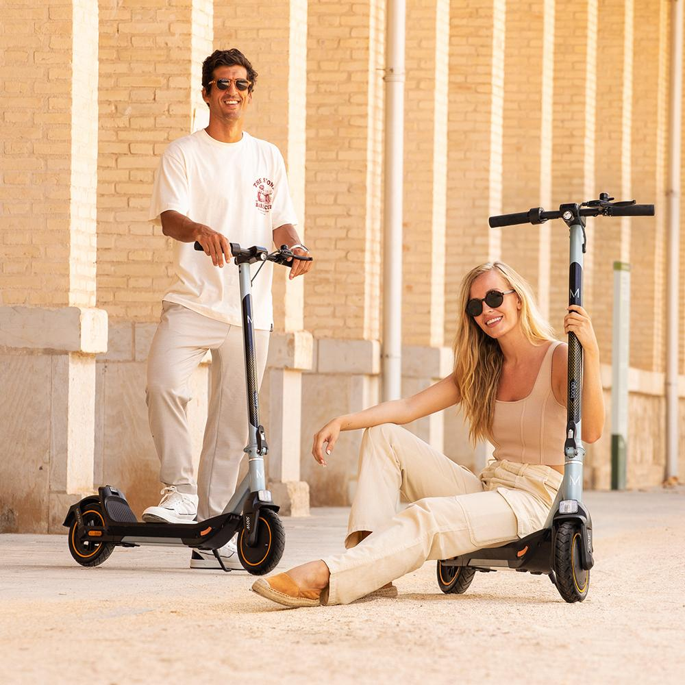 Un hombre y una mujer con patinetes, sonriendo. El hombre está de pie, la mujer sentada. Ropa beige, gafas de sol. Pared de ladrillo.