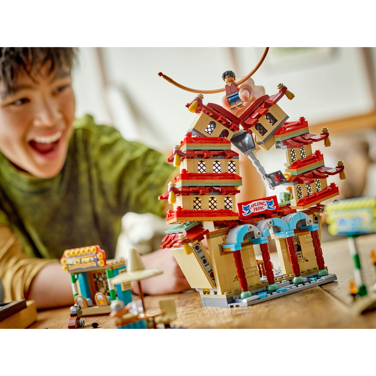 Un ragazzo sorridente guarda un set Lego, un edificio a più piani con tetti rossi e pareti beige. Una piccola figura è posta sopra.