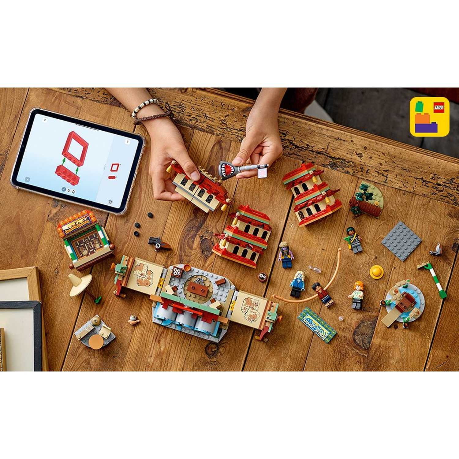 Mani che assemblano un set Lego su un tavolo di legno. I componenti includono edifici, mini-figure e un tablet che mostra le istruzioni.
