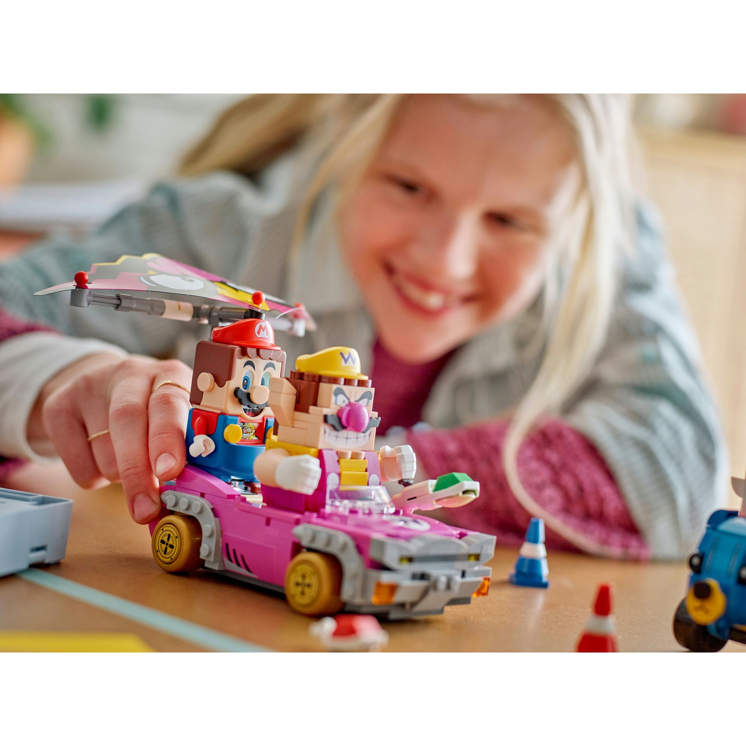 Una ragazza sorride a un'auto Lego rosa con le figure di Mario e Wario. Altri pezzi Lego sono visibili su un tavolo di legno.