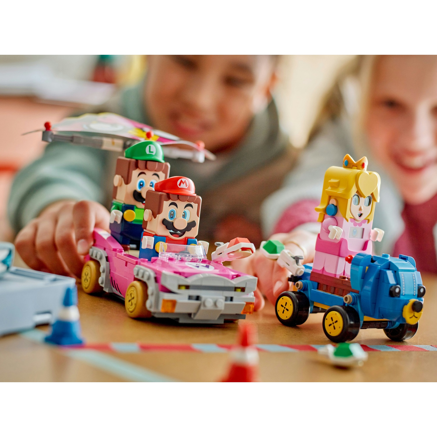 Lego Mario, Luigi e Peach in macchinine. Bambini sullo sfondo. Auto rosa, blu e grigie su un tavolo con una pista.