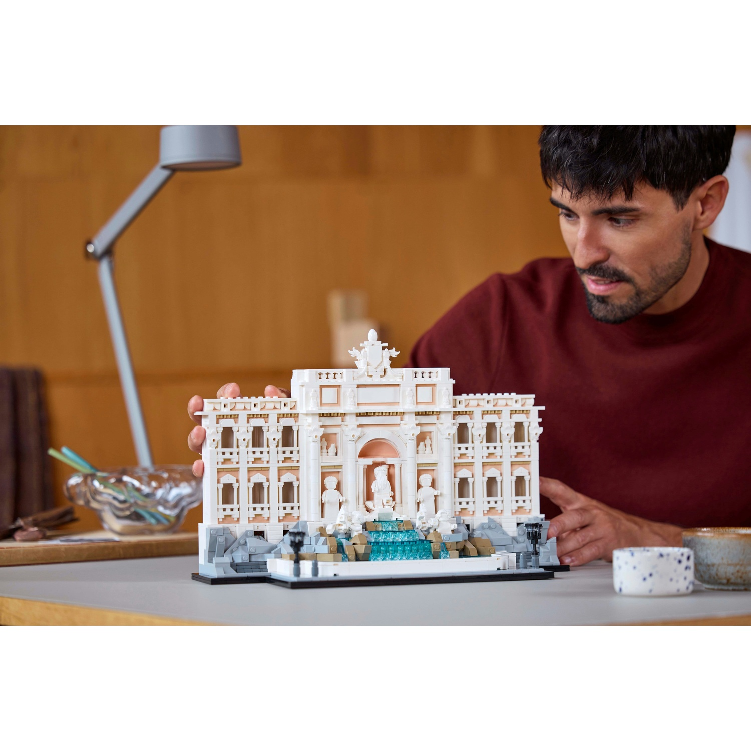 Un uomo tiene in mano un modello Lego di un edificio. Il modello è bianco con dettagli architettonici. L'uomo indossa una camicia bordeaux.