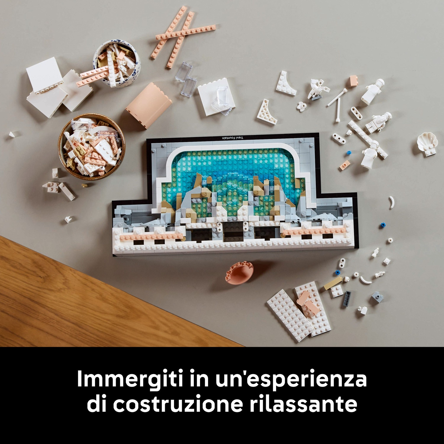 Un set Lego di una fontana in costruzione, con pezzi sparsi, ciotole di blocchi e la struttura parzialmente assemblata su una superficie grigia.