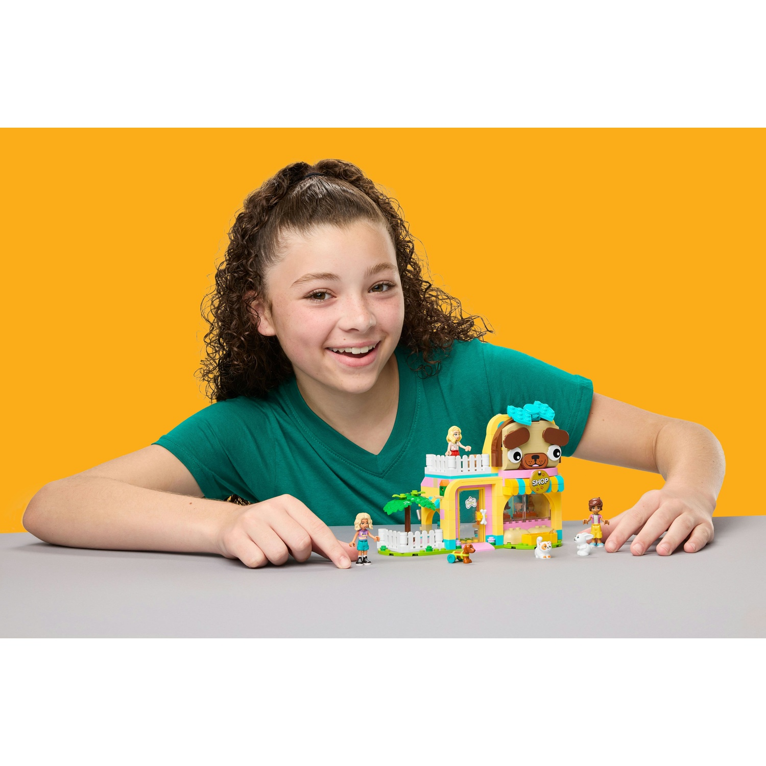 Una ragazza sorride alla fotocamera, appoggiata a un tavolo con un colorato set LEGO a tema cani. Il set ha minifigure e animali giocattolo.