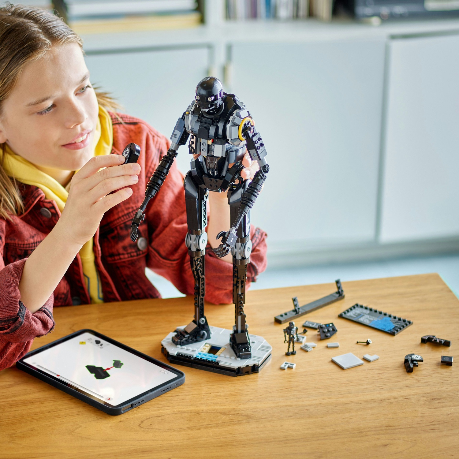 Una ragazza assembla un giocattolo robot nero. Un iPad mostra le istruzioni. Parti di robot e una piccola figura sono su un tavolo di legno.