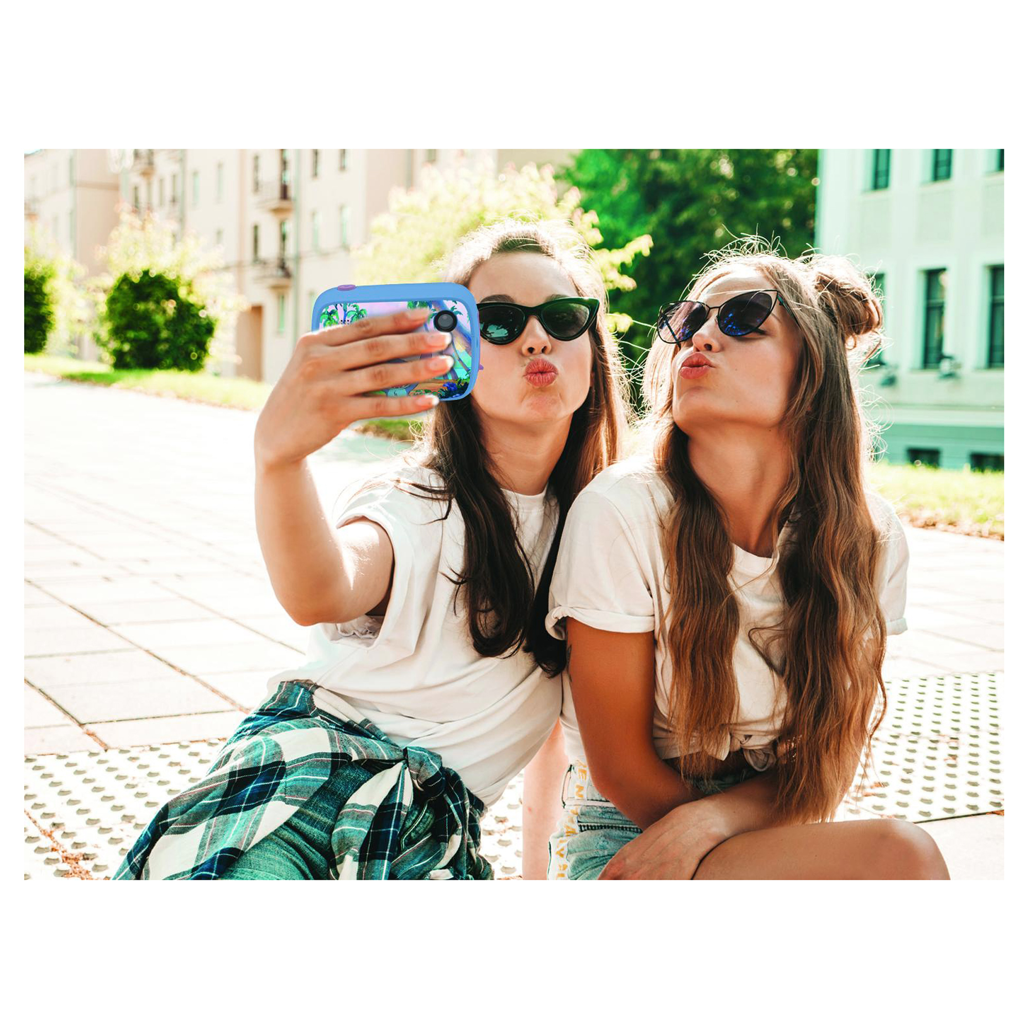 Dos mujeres jóvenes se hacen un selfie, frunciendo los labios. Una sostiene el teléfono, lleva gafas de sol. El fondo tiene árboles y edificios.