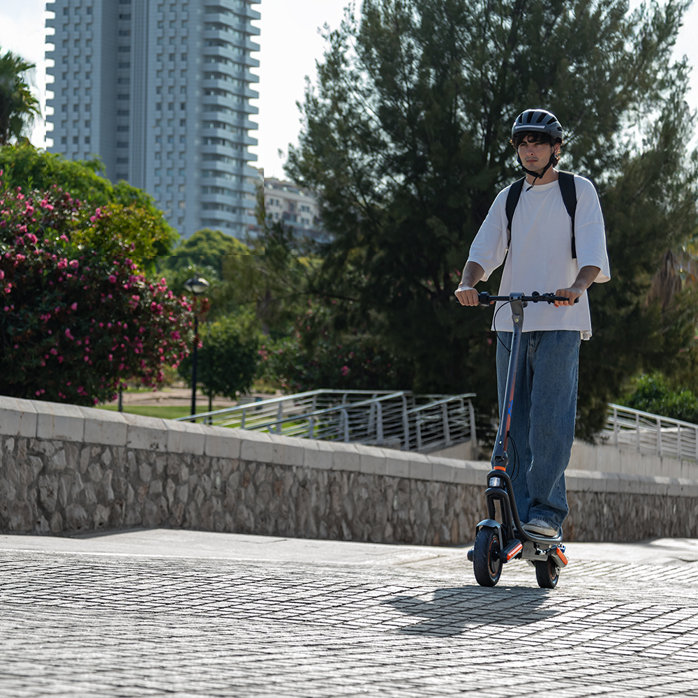 Un hombre con casco monta un patinete eléctrico. Edificio alto y árboles al fondo. Flores rosas a lo largo de un muro.