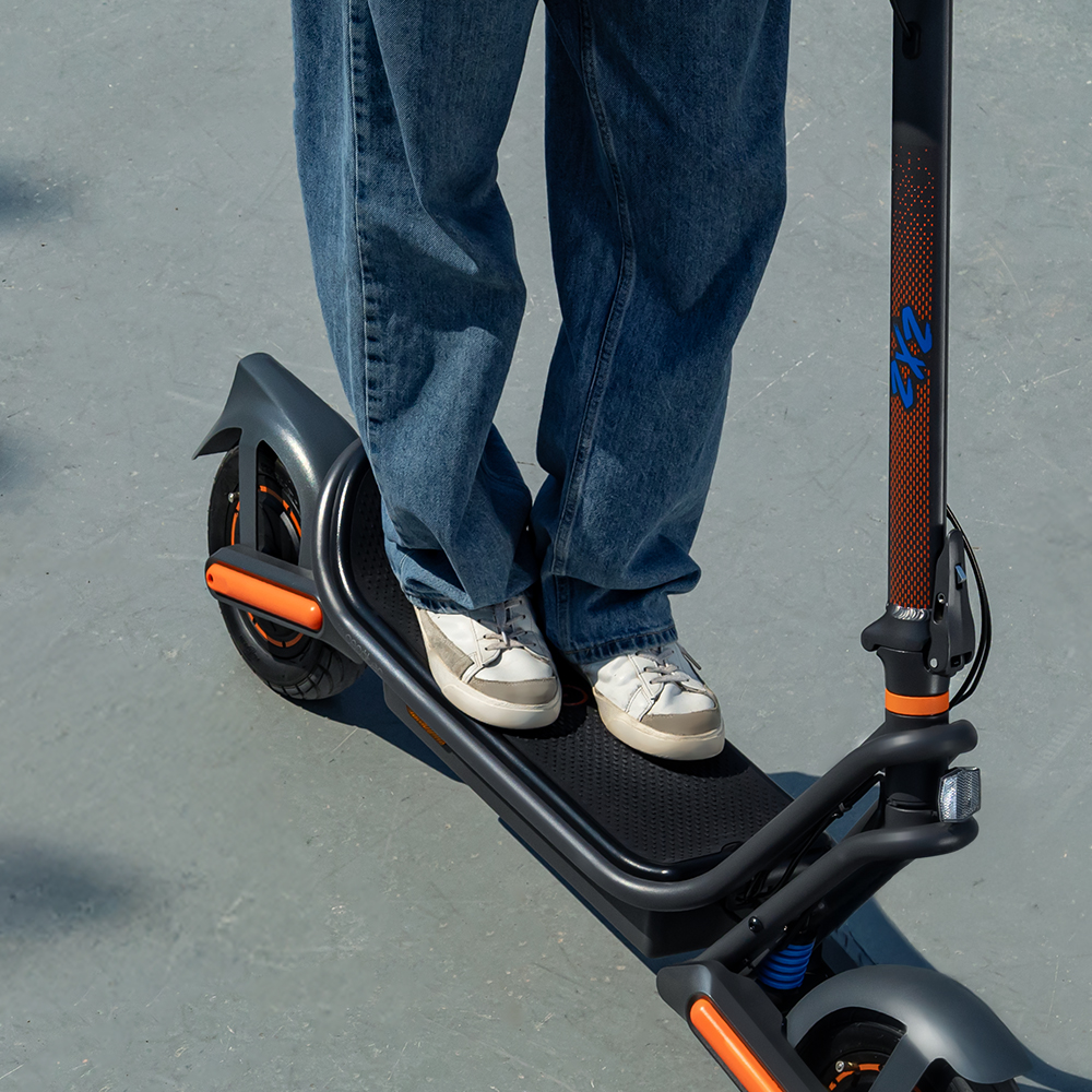 Una persona está de pie sobre un scooter. Lleva vaqueros y zapatillas blancas. El scooter es gris y negro con detalles naranjas.