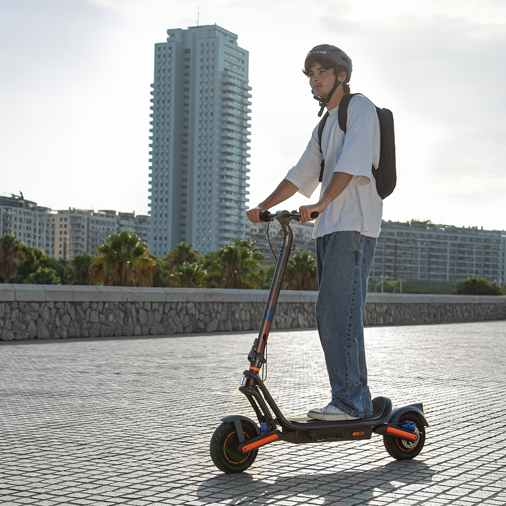 Una persona en un patinete eléctrico. Llevan casco y mochila. Edificios y un muro de piedra al fondo.