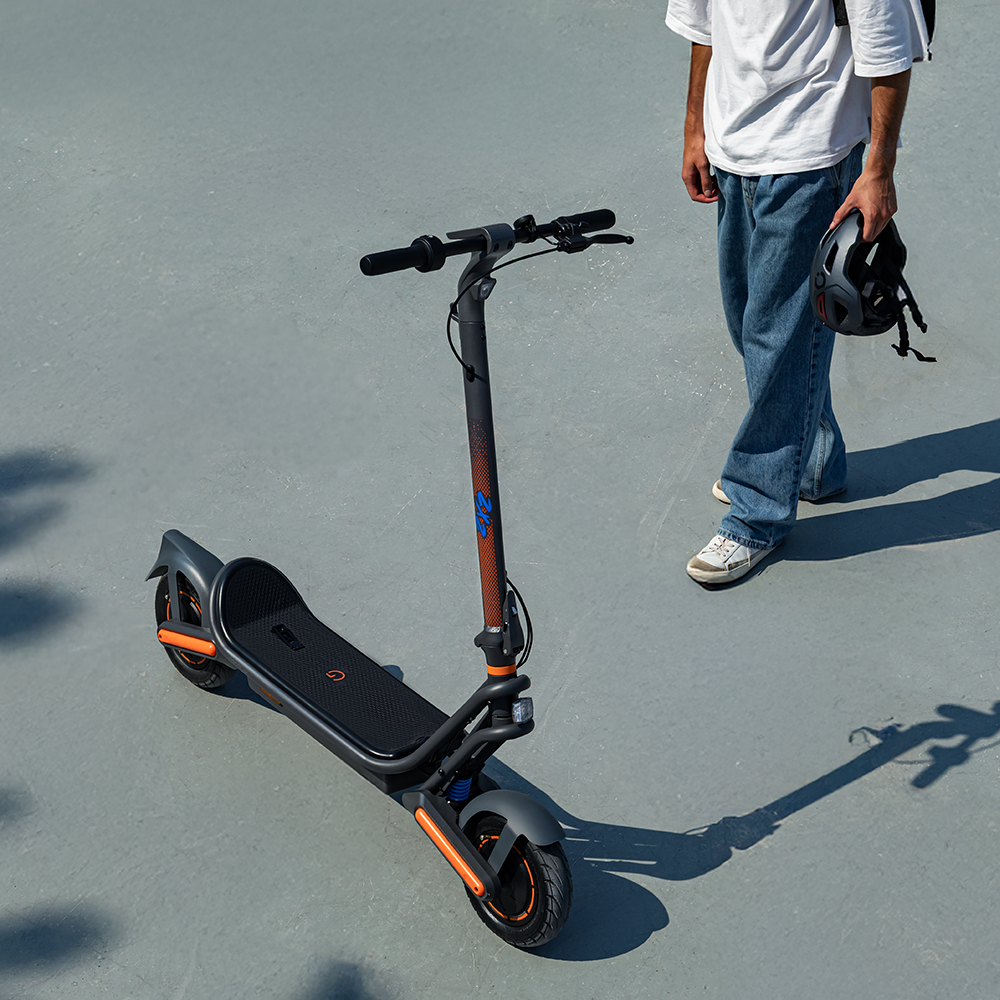 Una persona está de pie con un patinete eléctrico. El patinete es negro y naranja. La persona sostiene un casco, usa jeans y una camisa blanca.