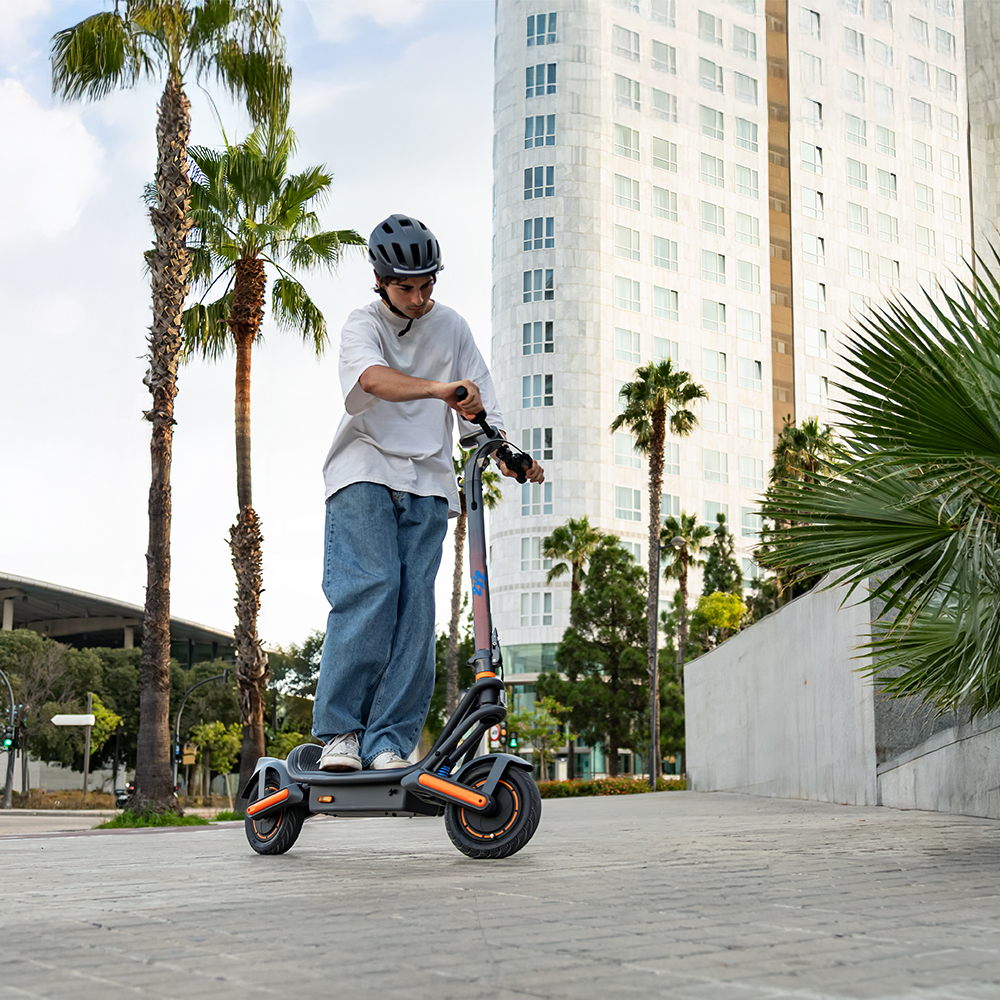 Una persona monta un patinete eléctrico en una acera. Hay palmeras y un edificio al fondo. La persona lleva un casco.