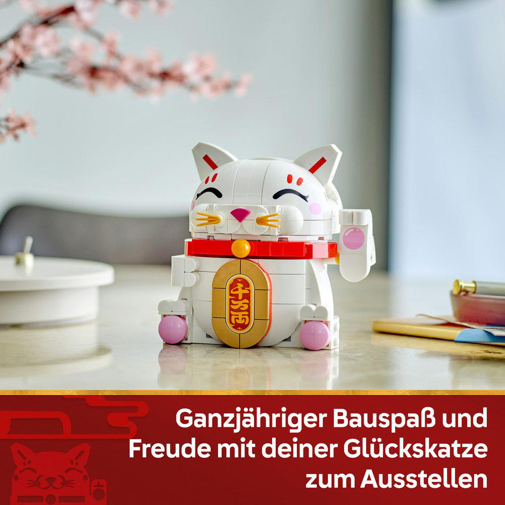 Ein weißes Glückskatzenspielzeug mit roten Akzenten und einem goldenen Anhänger. Es steht auf einem Tisch. Im Hintergrund Kirschblüten und Text.