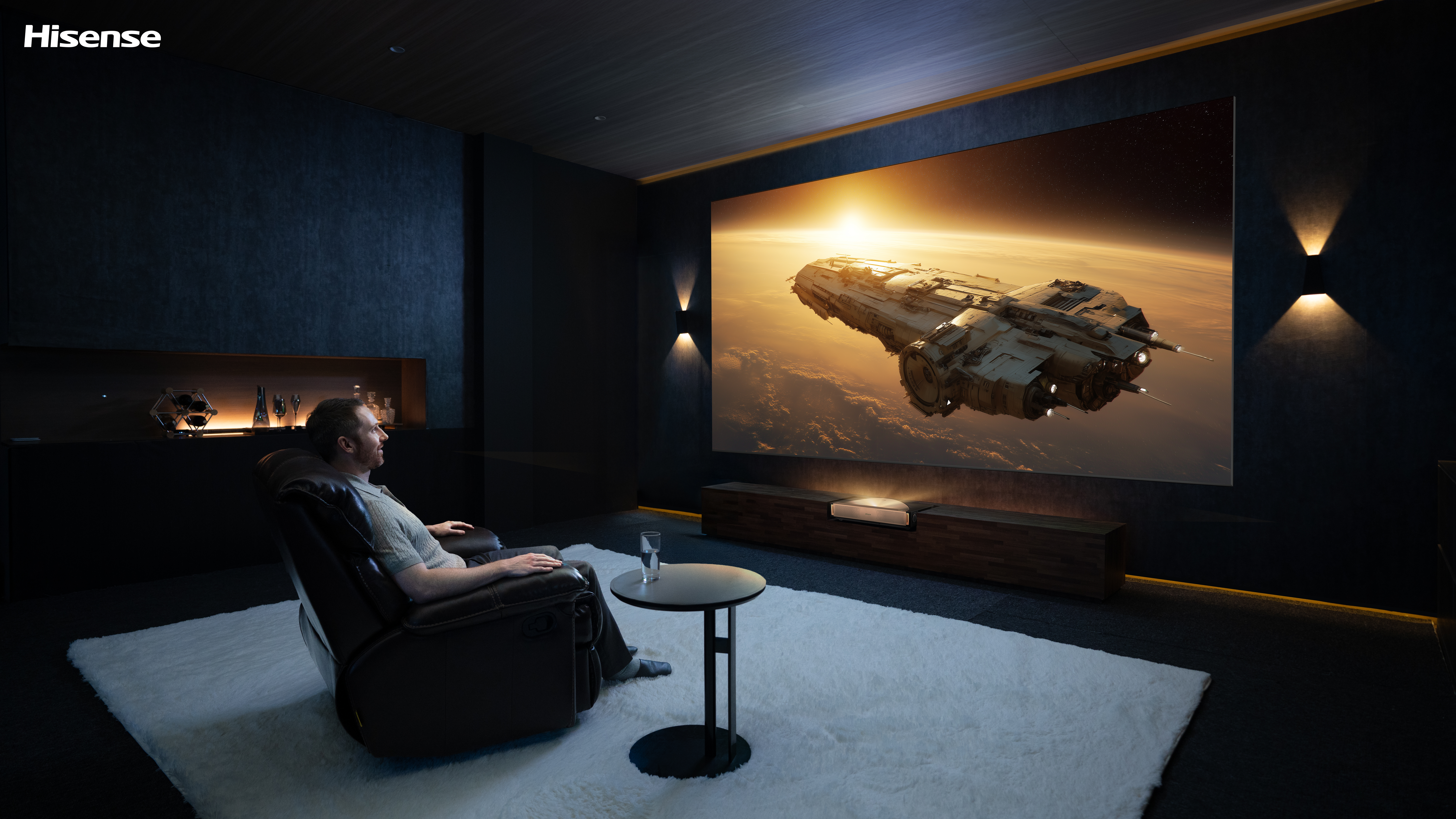 Mężczyzna ogląda statek kosmiczny na dużym ekranie w ciemnym pokoju. Siedzi w fotelu z stolikiem i szklanką wody.
