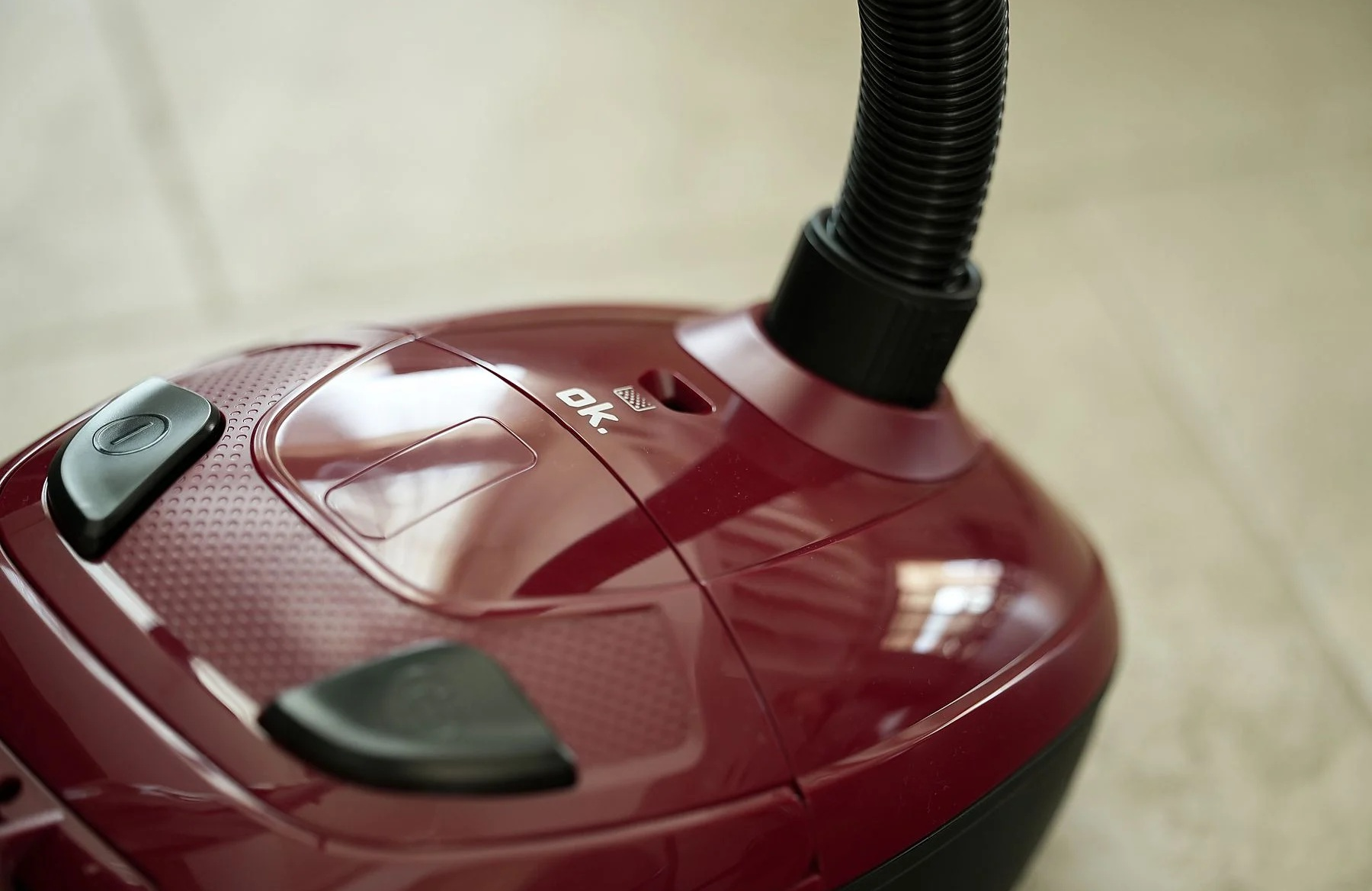 Zbliżenie na bordowy odkurzacz z czarnym wężem. Zawiera przyciski, logo 'OK' i teksturowaną powierzchnię.