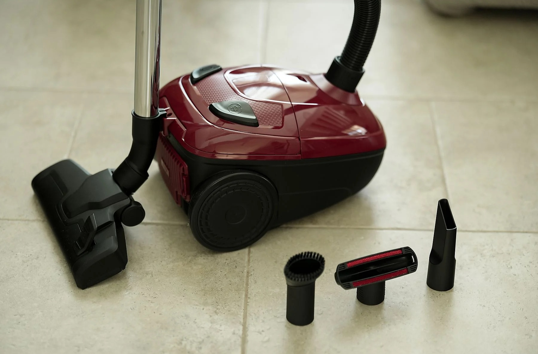 Bordowy odkurzacz z czarnymi nasadkami stoi na kafelkach. Dodatkowe nasadki leżą obok.