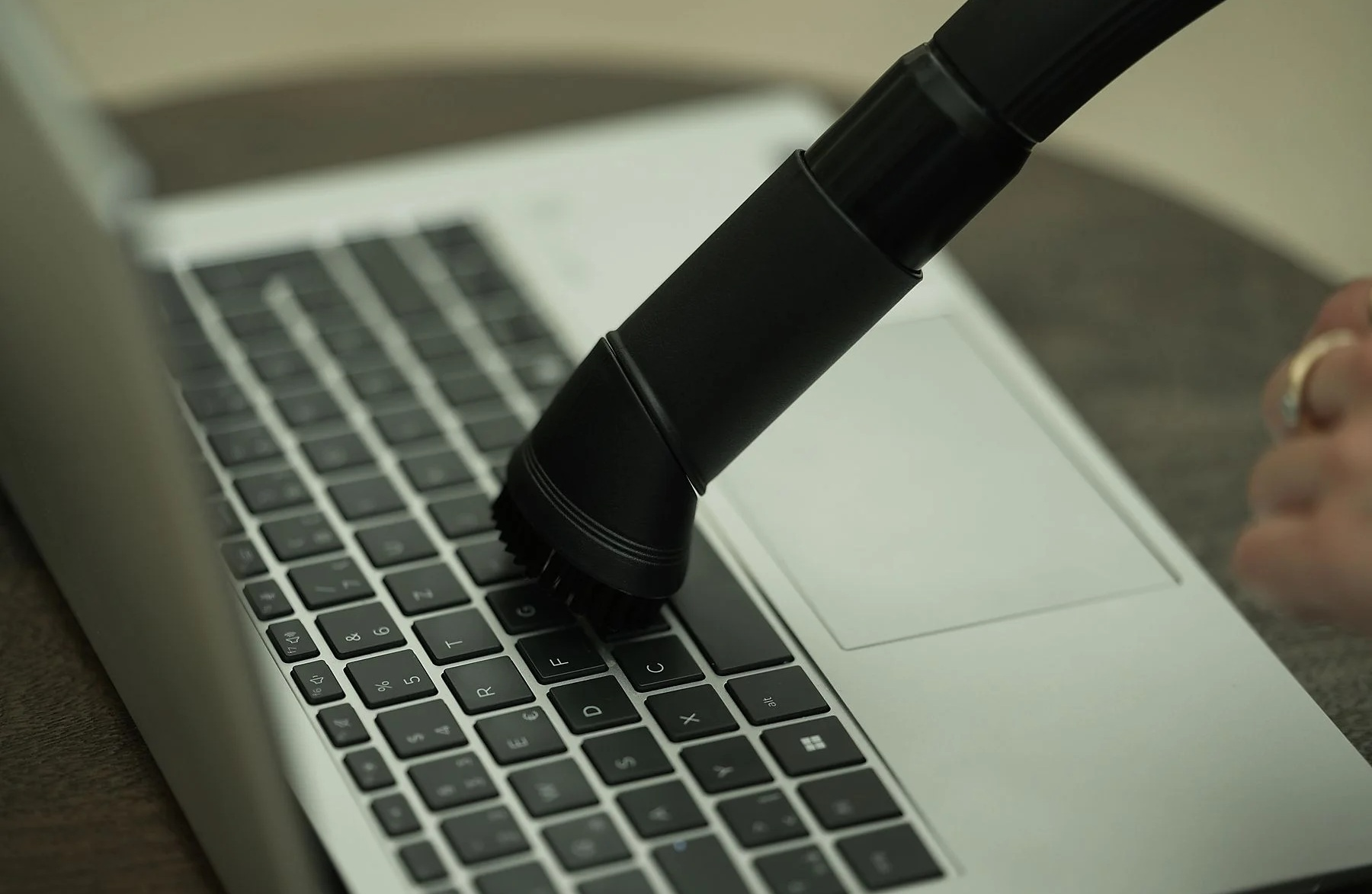 Czarna głowica odkurzacza czyści klawiaturę laptopa. Laptop jest otwarty na stole. Ręka z pierścieniami po prawej stronie.