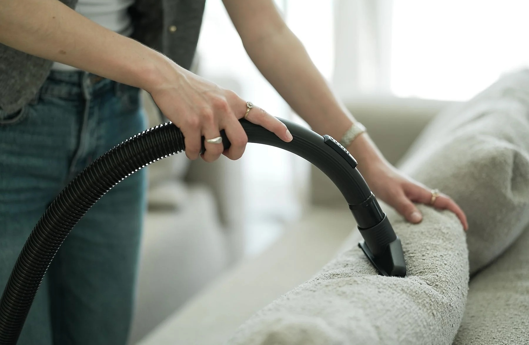 Osoba odkurza jasną sofę czarnym odkurzaczem. Osoba ma na sobie dżinsy i szarą kamizelkę.