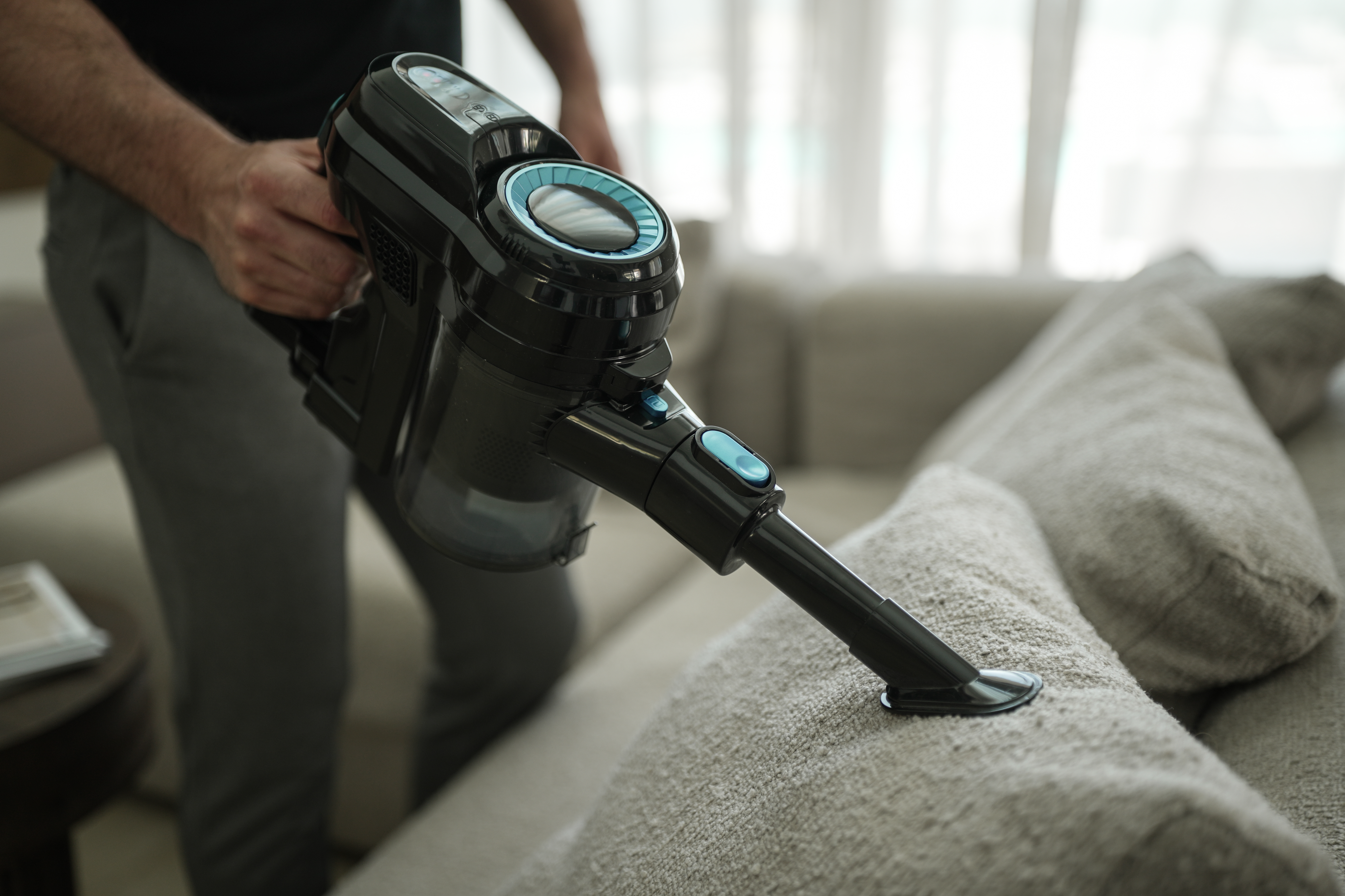 Osoba odkurza sofę czarno-niebieskim odkurzaczem ręcznym. Sofa jest beżowa.