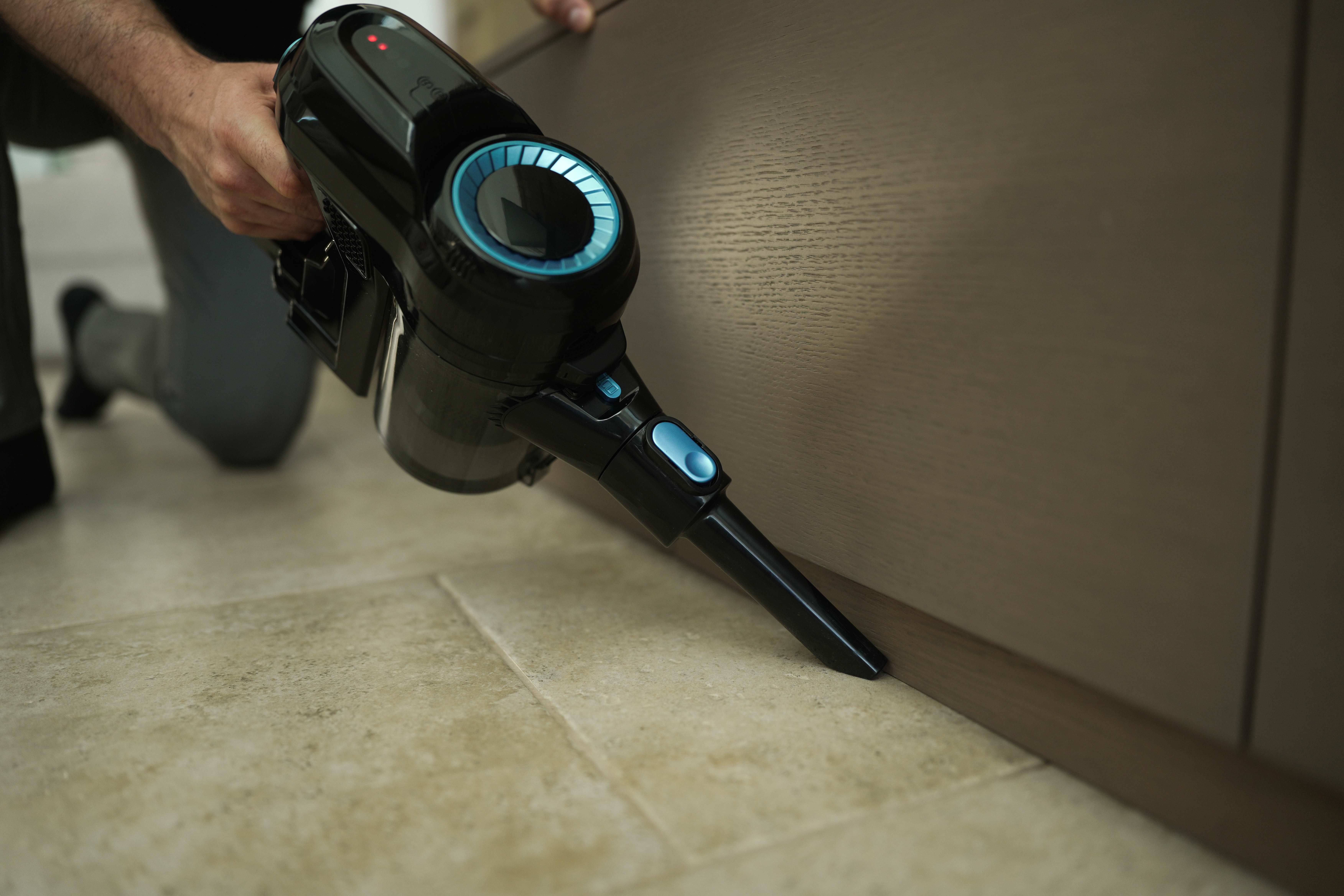 Osoba używa czarno-niebieskiego odkurzacza ręcznego na kafelkach w pobliżu szafki.