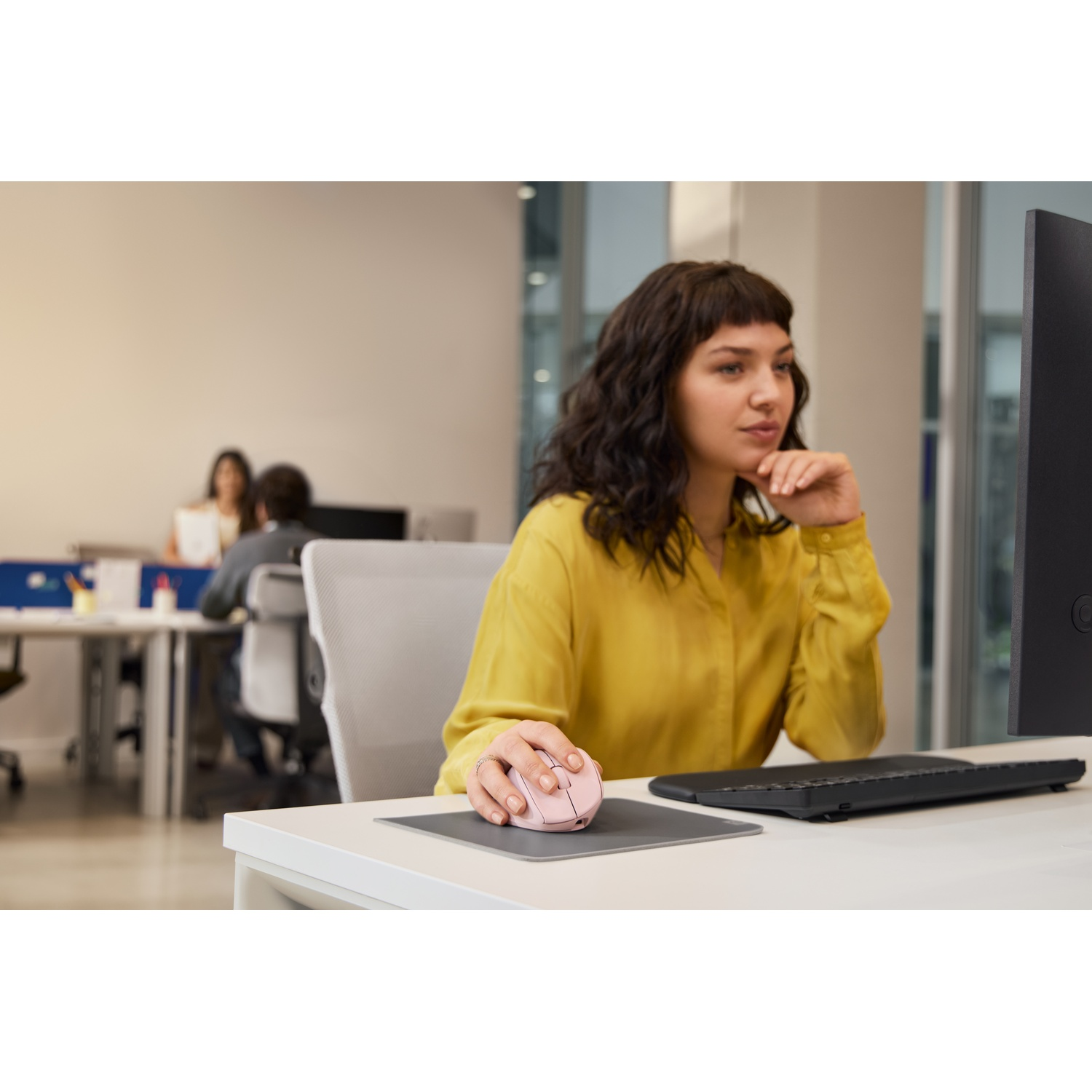 Une femme en chemise jaune utilise une souris rose à un bureau. Il y a un ordinateur et d'autres personnes en arrière-plan.