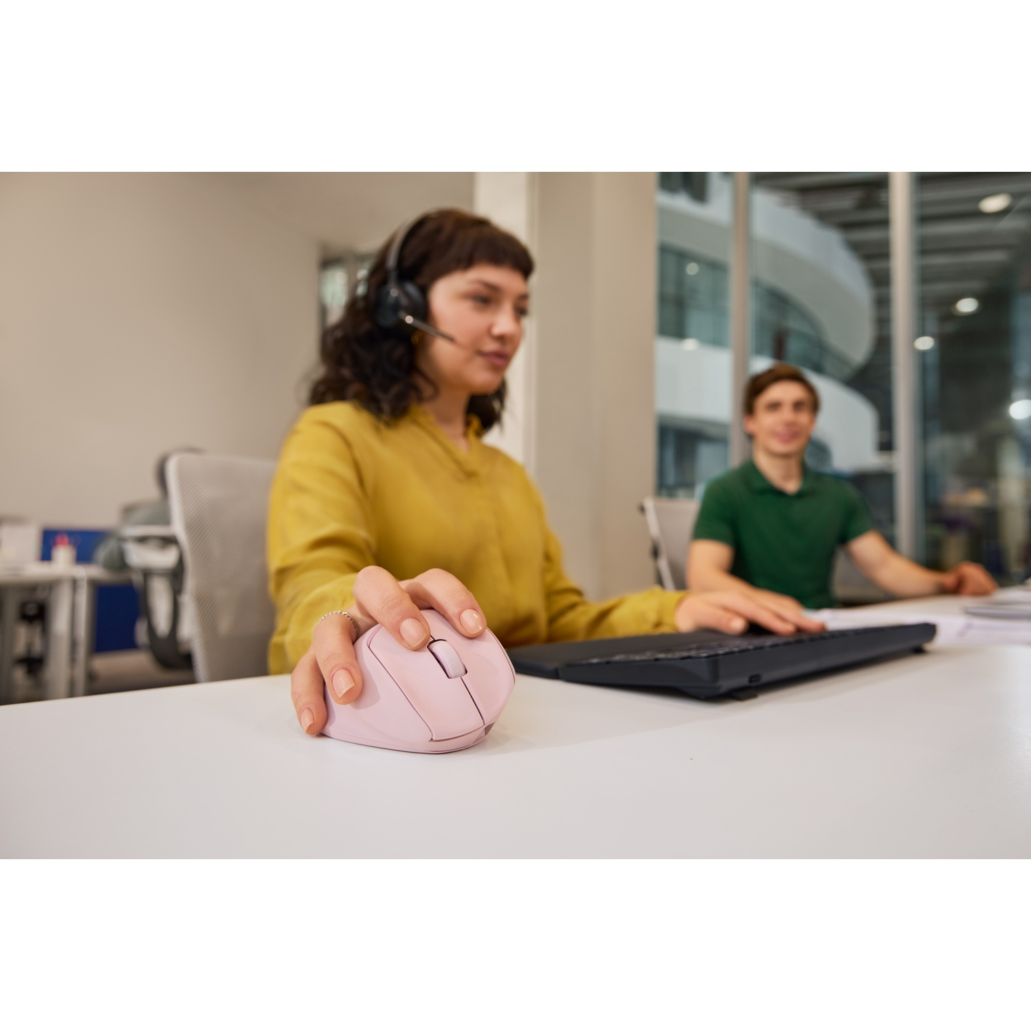Une femme avec un casque utilise une souris rose. Un homme tape sur un clavier derrière elle. Tous deux sont à un bureau blanc dans un bureau.
