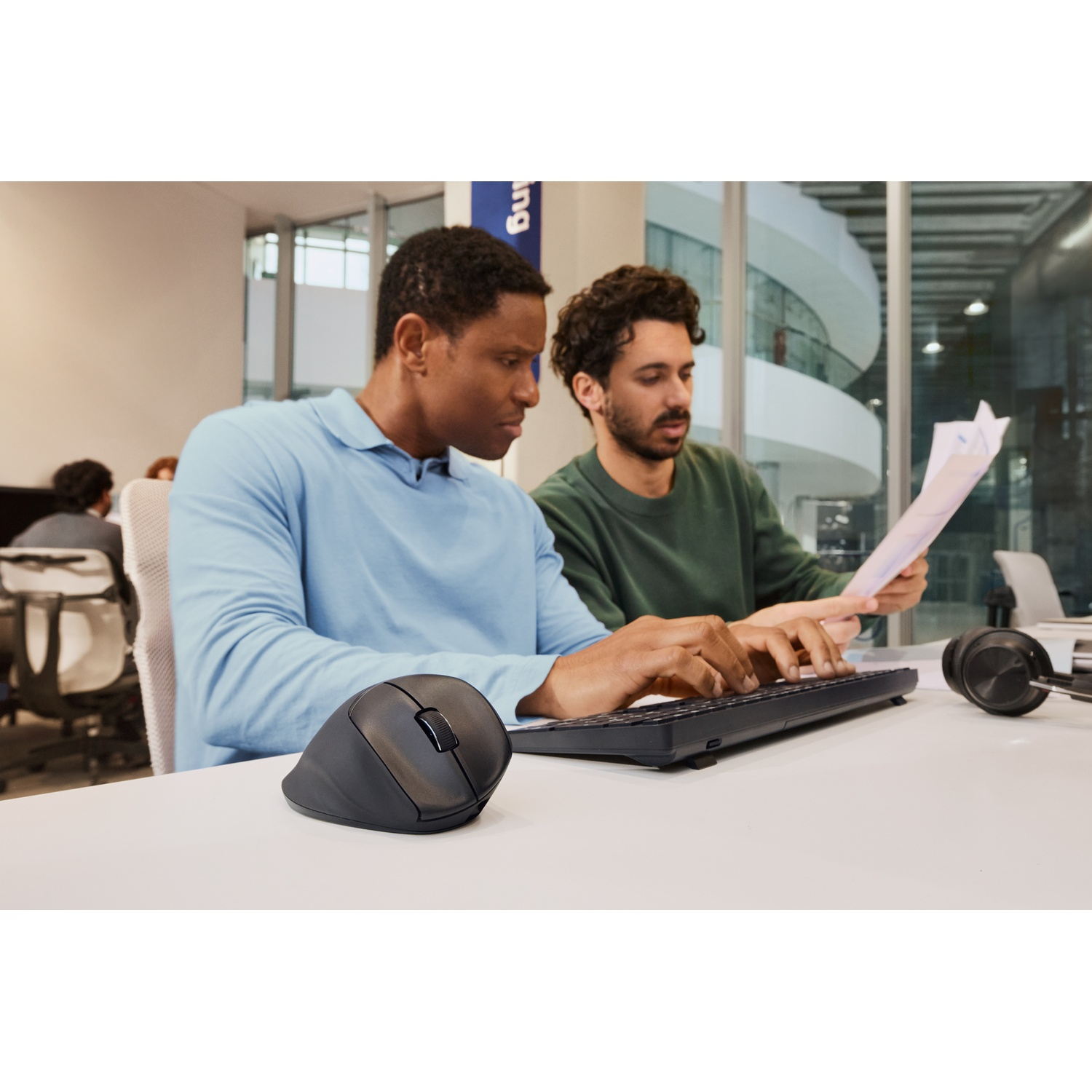 Deux hommes à un bureau, l'un tapant sur un clavier, l'autre tenant des papiers. Une souris noire et un casque sont visibles.