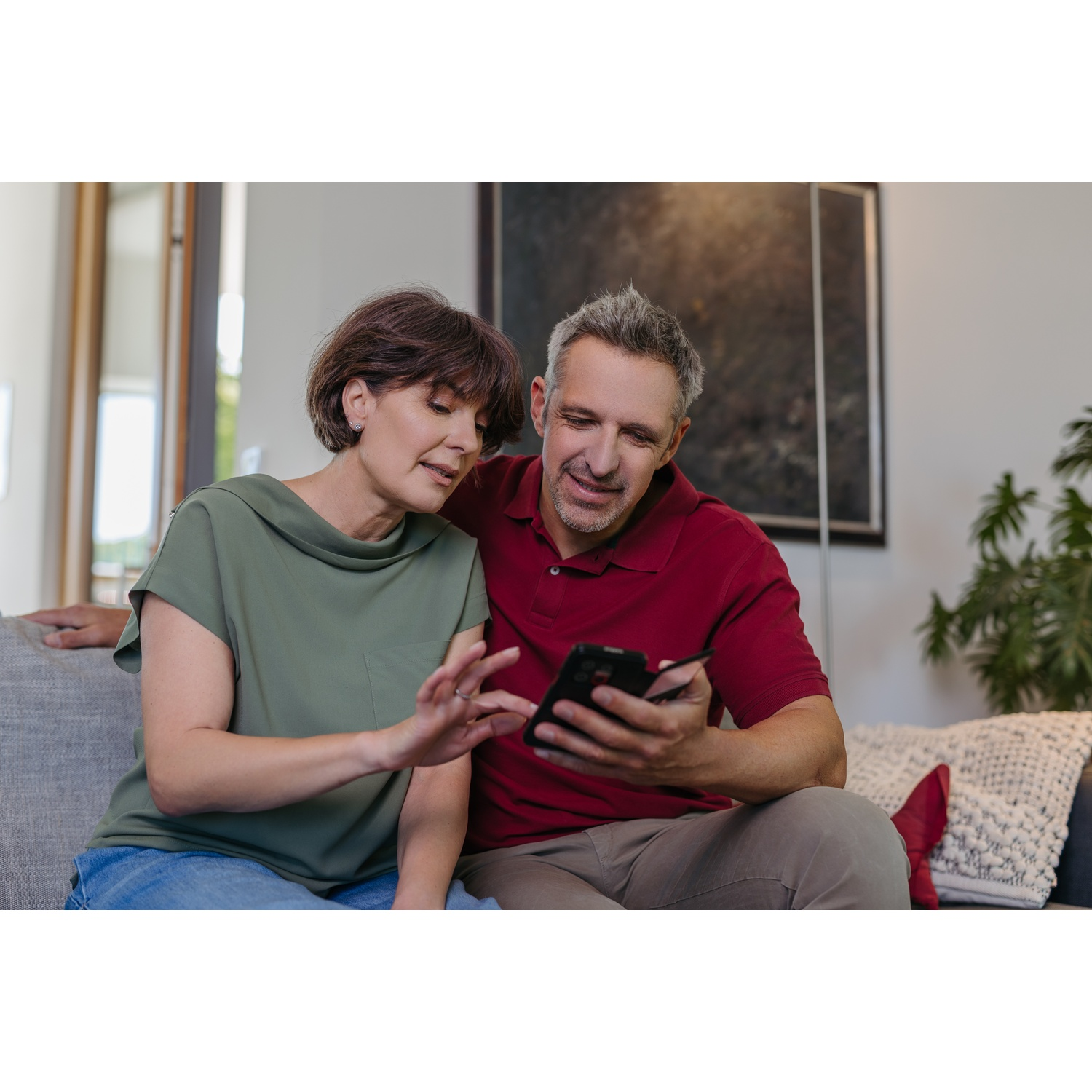 Een stel zit op een bank en bekijkt een telefoon. De vrouw draagt een groen shirt, de man een rood.