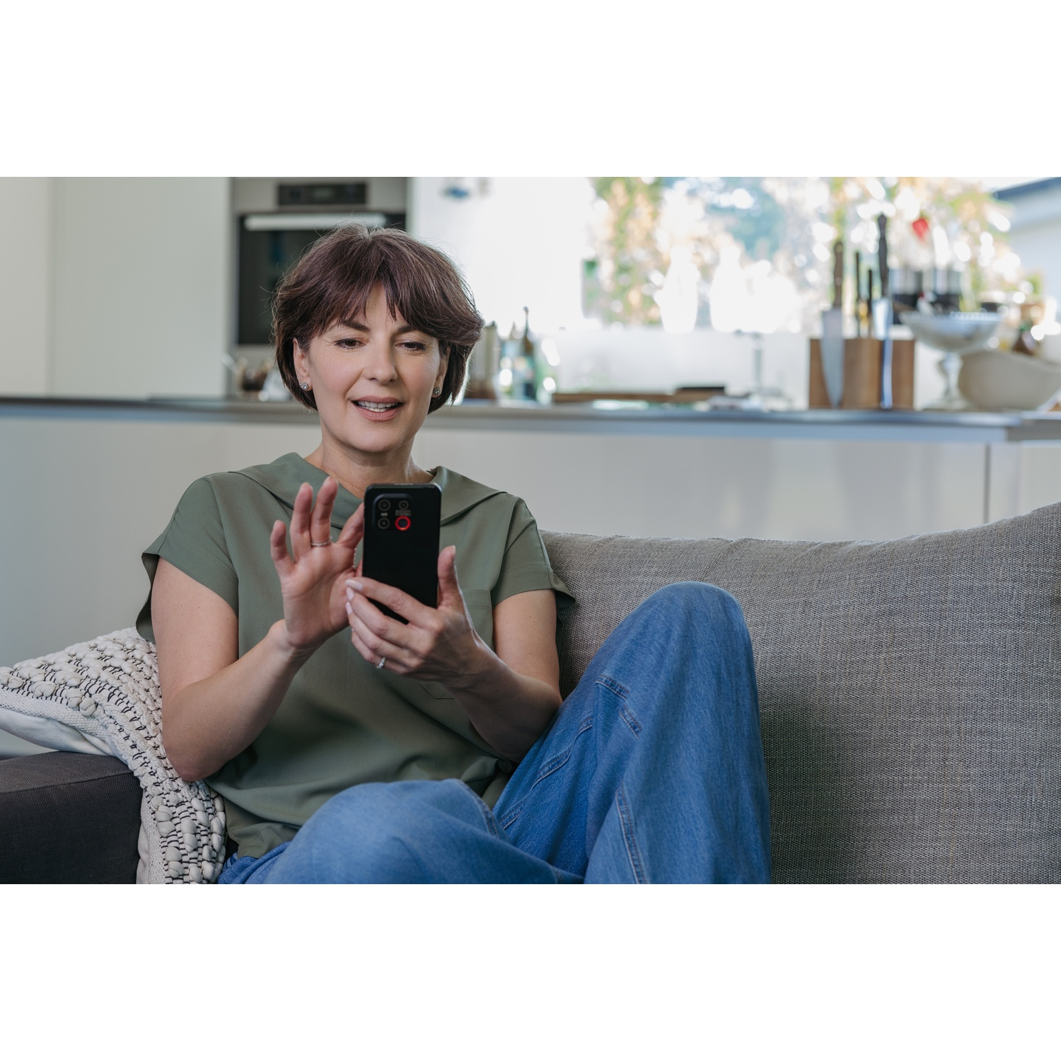 Vrouw op een bank met een telefoon. Ze draagt een groen shirt en spijkerbroek. Achtergrond toont een keuken.