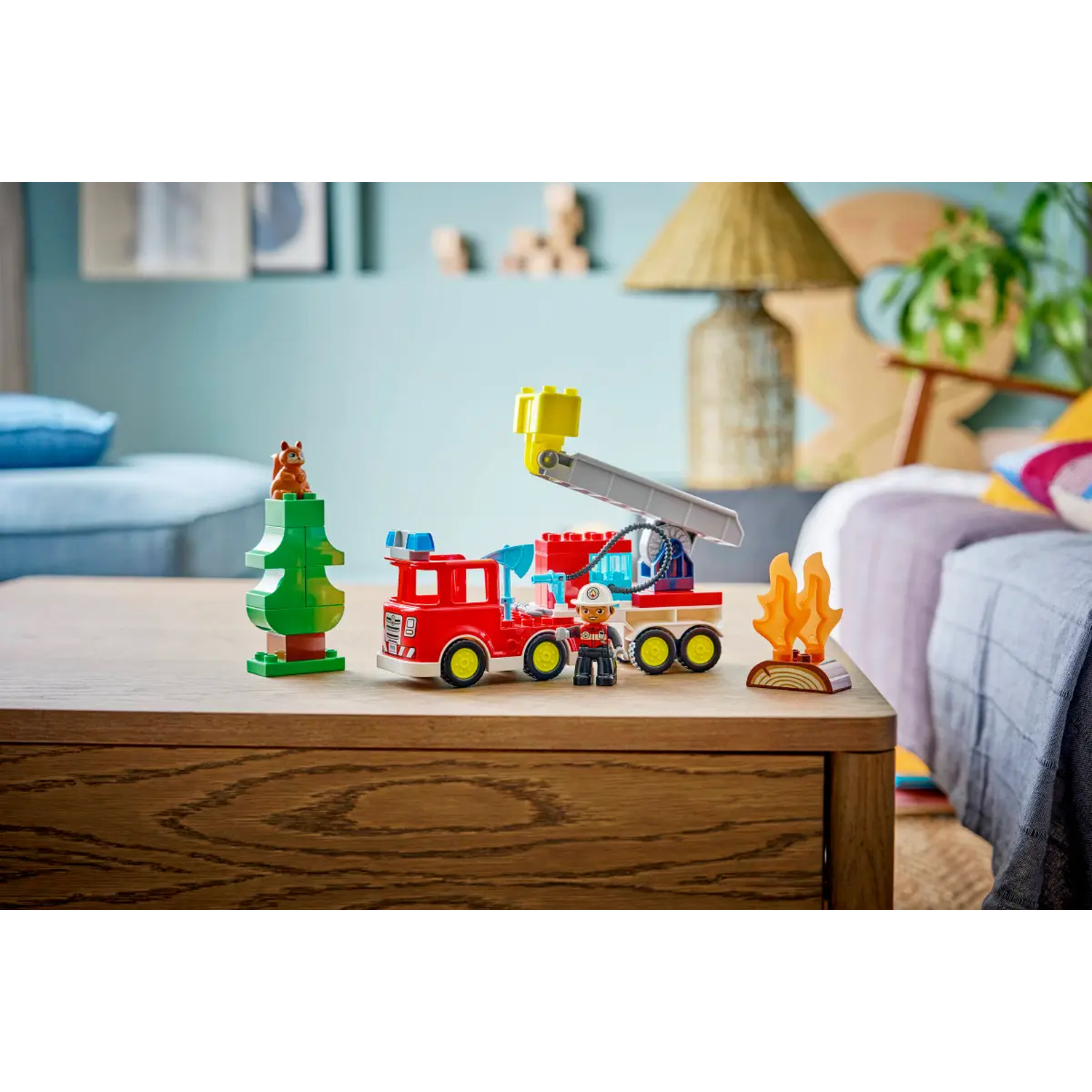 A Lego fire truck, tree with squirrel, and a fire scene are on a wooden table. A firefighter figure stands nearby.