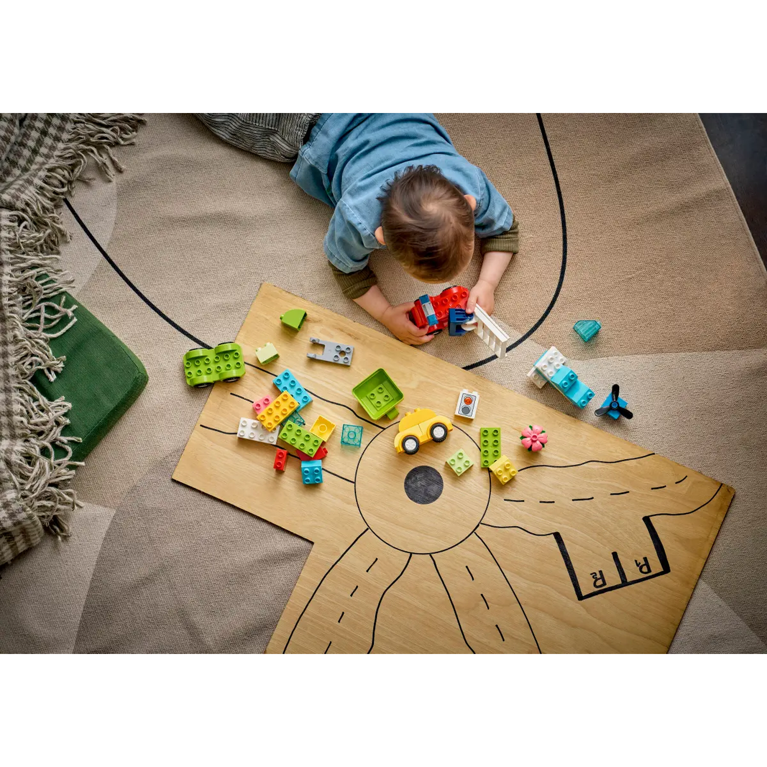 A child plays with building blocks on a wooden board with road markings. A green cushion and a blanket are in the background.