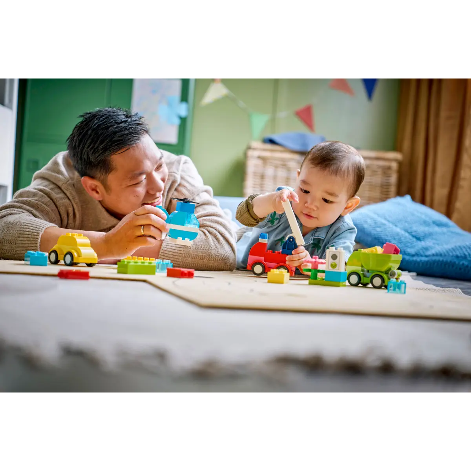 A man and a baby playing with colorful toy blocks. They are on a light surface with toys. Background has a basket and wall.