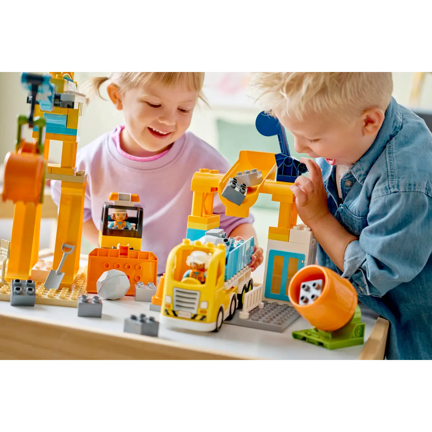 Two children playing with a construction toy set. The set includes a truck, crane, and other building elements. They are smiling.