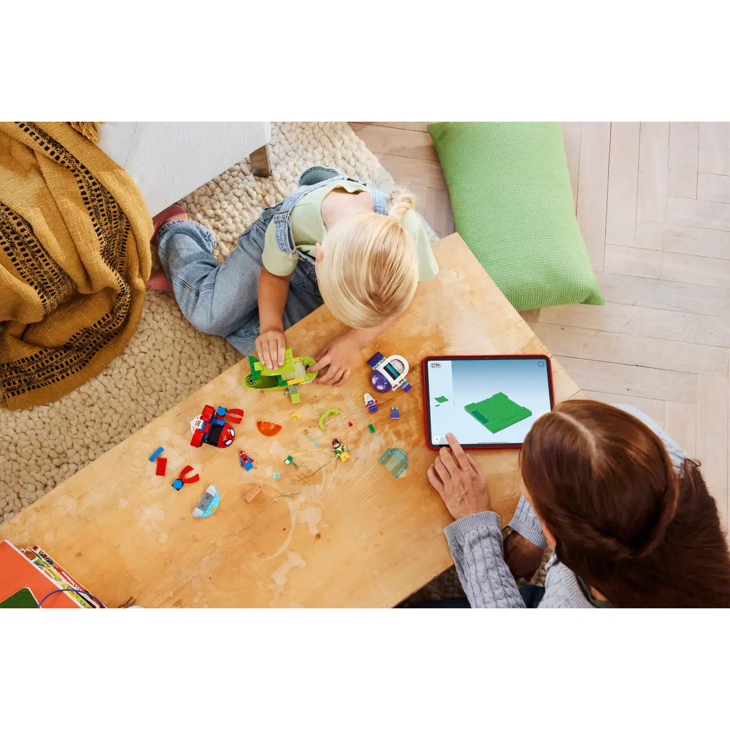 A child and an adult are playing with toys on a wooden table. The child is building with blocks, and the adult is using a tablet.