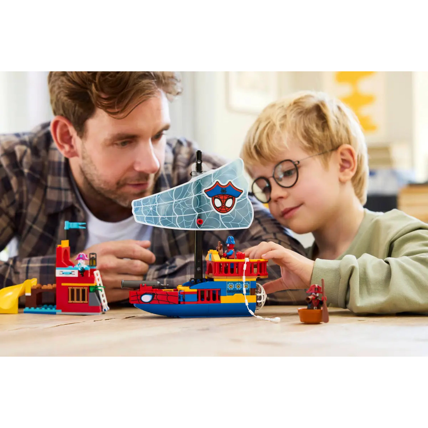 A man and a boy are playing with a Spider-Man LEGO set, featuring a boat and a building, on a wooden table.