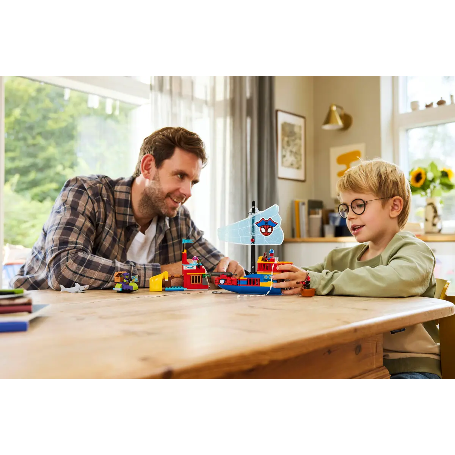 A man and a boy play with toy boats on a wooden table. The boy wears glasses. Sunlight streams in from a window.