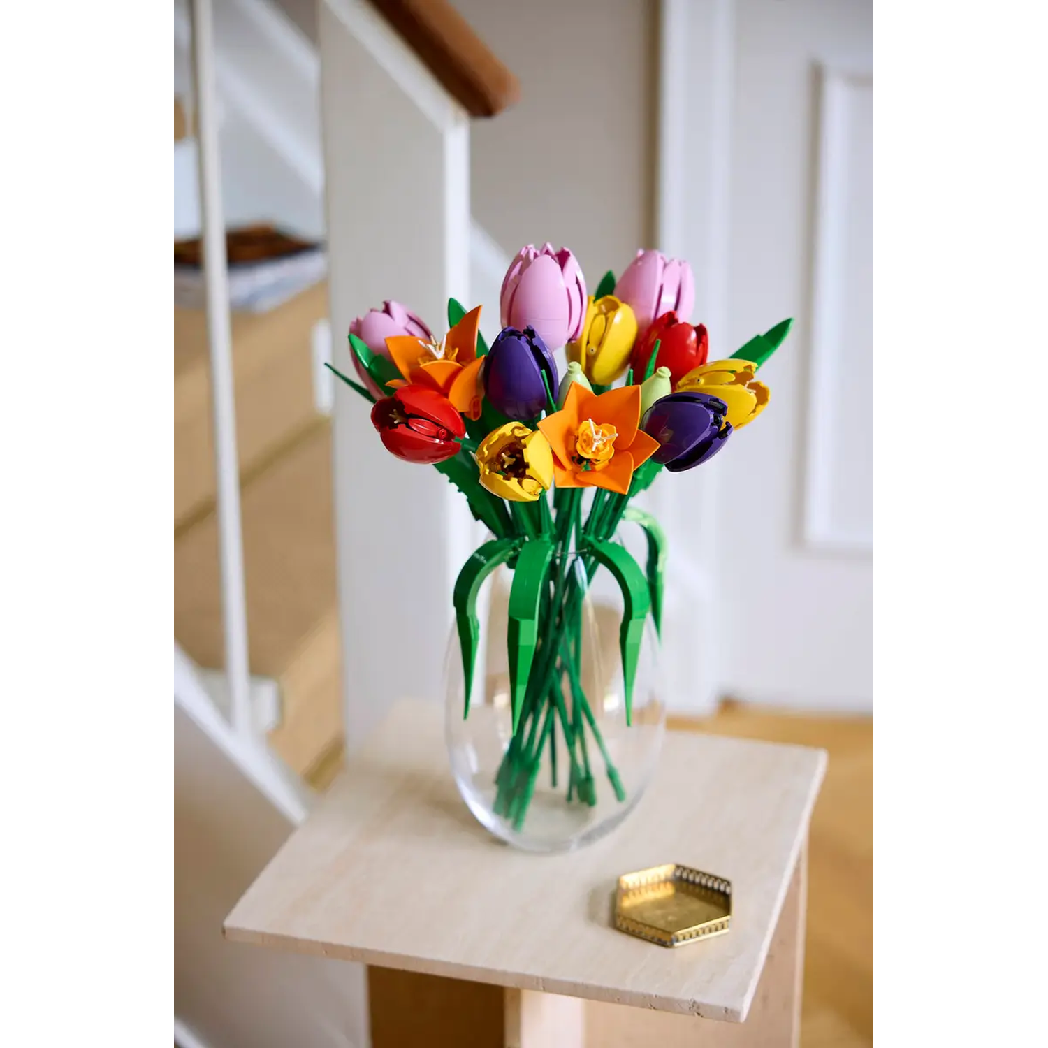 Een boeket kleurrijke Lego-tulpen in een doorzichtige vaas op een houten tafel. Een kleine gouden schaal staat naast de vaas. Trappen op de achtergrond.
